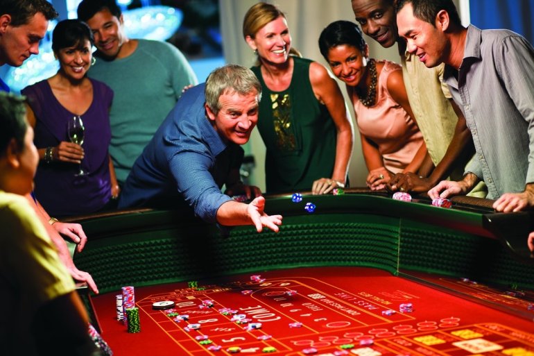 Игрок бросает кости на красный стол для крэпса, а люди разных национальностей наблюдают за его действем